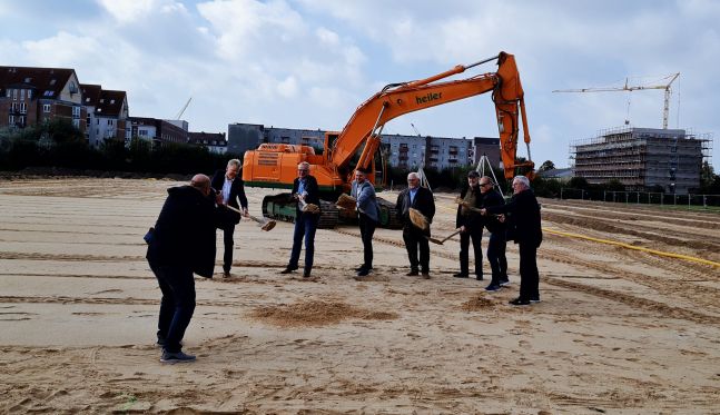 Auf einer großen Baustelle. Mehrere Männer mit Schaufeln in der Hand schaufeln symbolisch Sand. Im Hintergrund steht ein Bagger.