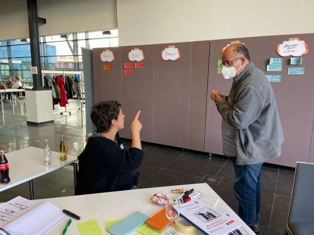 Ein Mann und eine Frau schauen auf die Arbeitsergebnisse einer Arbeitsgruppe, die auf Karten an eine Wand geklebt wurden