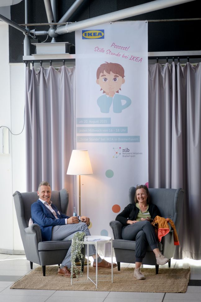 Zwei graue Ohrensessel auf einem Sisalteppich, eine Stehlampe mit einem weißen Schirm und ein kleiner weißer Tablettisch mit einer Blume. Ein Mann und eine Frau sitzen in den Sesseln. Ein Banner im Hintergrund weist auf die Stille Stunde bei Ikea hin.