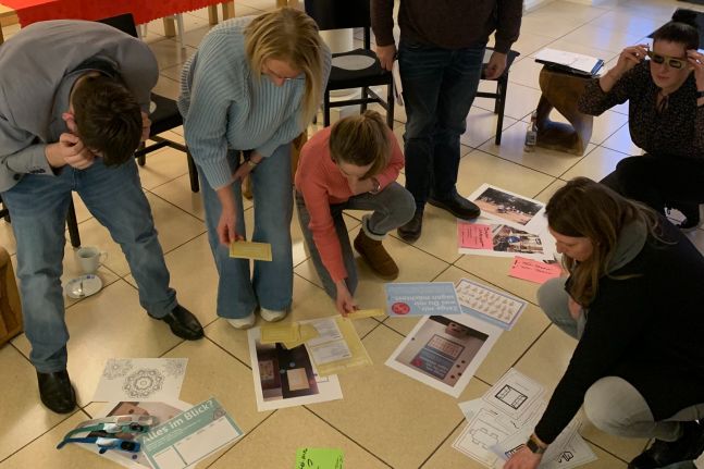 Mitglieder der Arbeitsgruppe bearbeiten Bilder und Bastelmaterial auf dem Boden.
