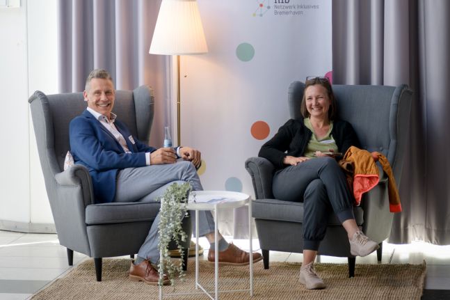Zwei graue Ohrensessel auf einem Sisalteppich, eine Stehlampe mit einem weißen Schirm und ein kleiner weißer Tablettisch mit einer Blume. Ein Mann und eine Frau sitzen in den Sesseln. Ein Banner im Hintergrund weist auf die Stille Stunde bei Ikea hin.