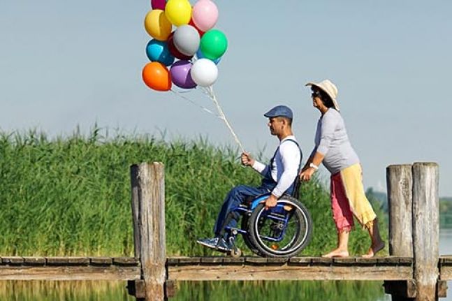 Mann im Rollstuhl hält eine große Traube mit Luftballons in die Luft, er wird von einer Frau über einen Steg geschoben